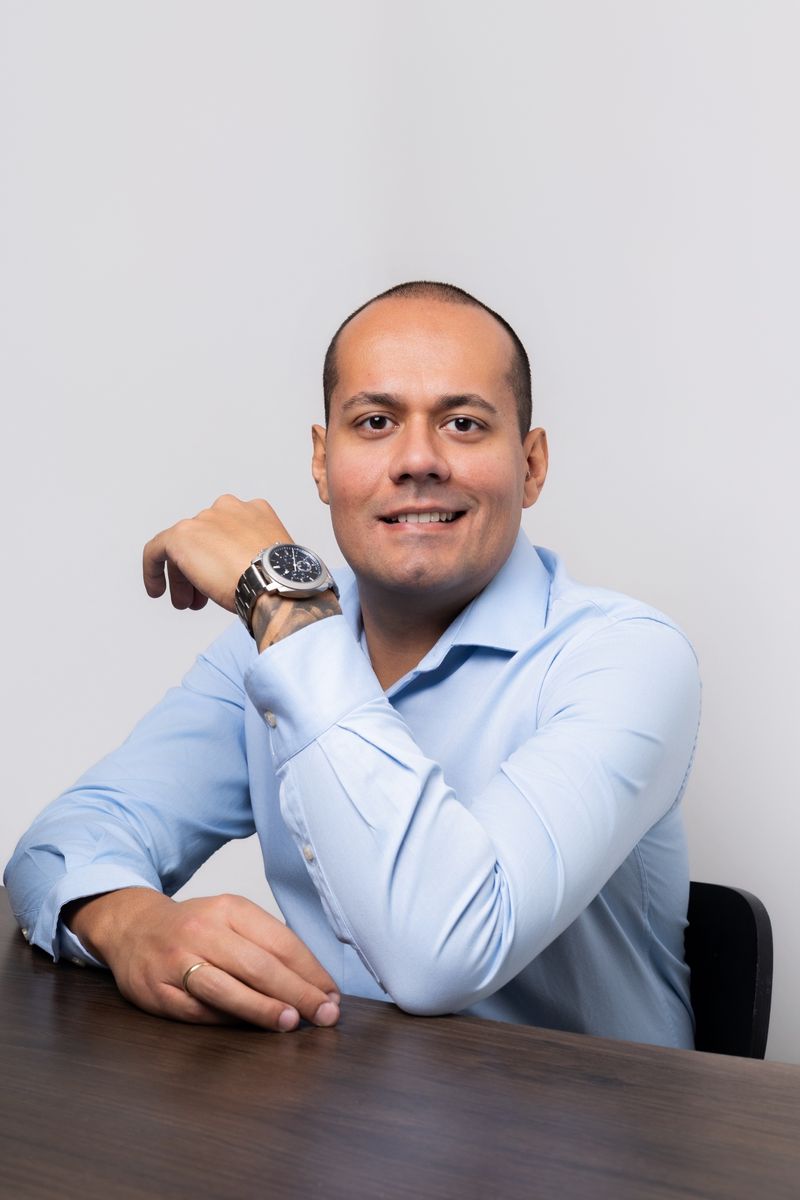 Matheus Marques sorrindo, de camisa azul, apoiado em uma mesa